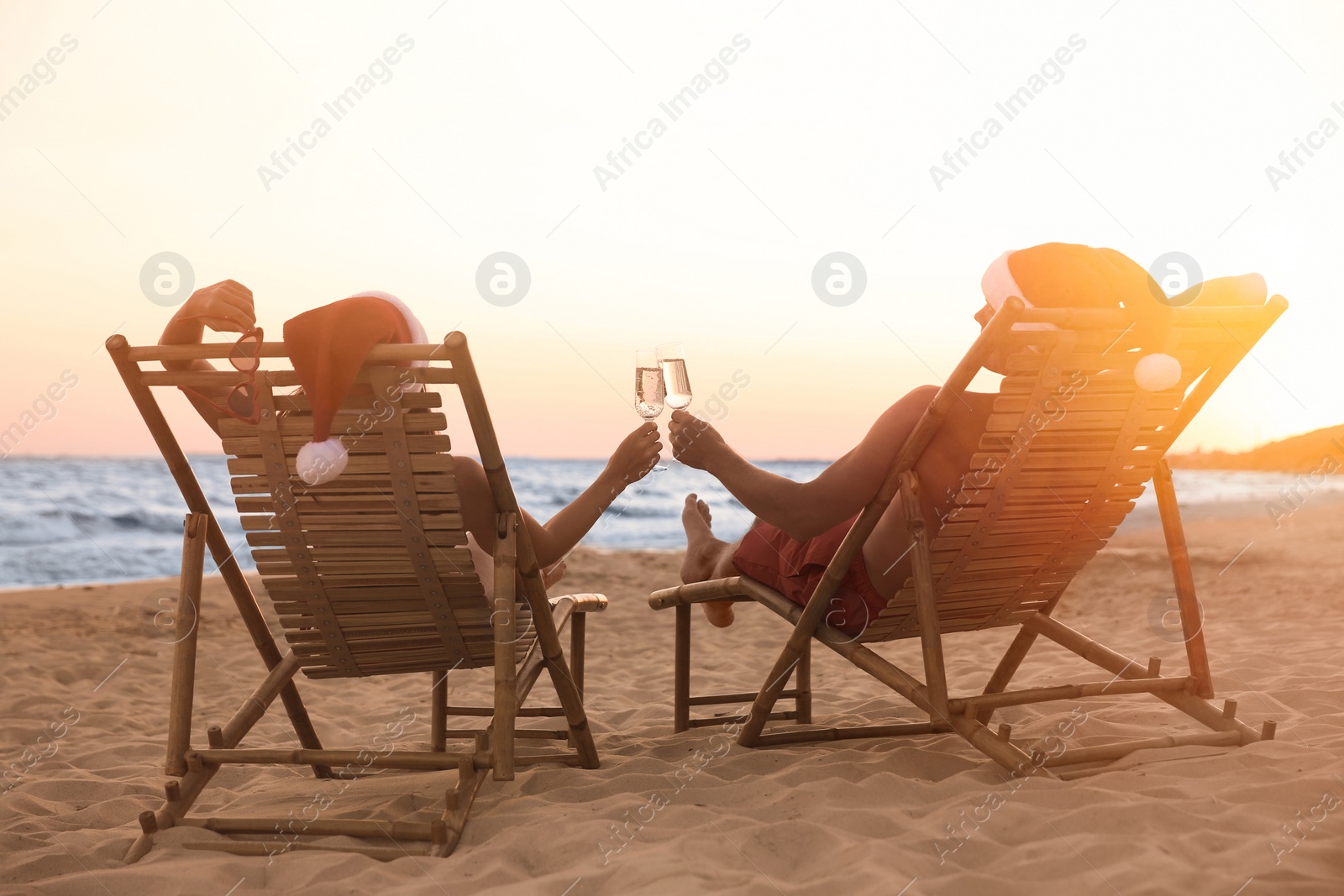 Photo of Lovely couple wearing Santa hats and drinking champagne together on beach. Christmas vacation