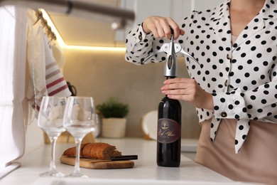 Photo of Romantic dinner. Woman opening wine bottle with corkscrew at countertop in kitchen, closeup