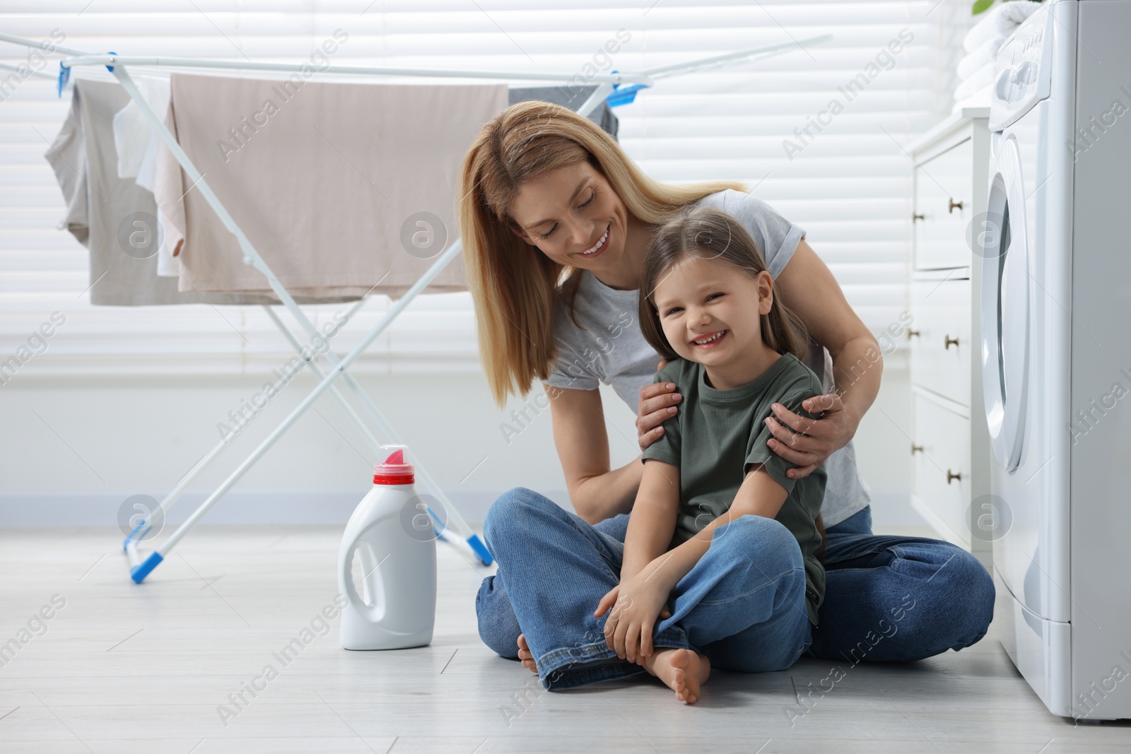 Photo of Mother and daughter sitting on floor near washing machine and fabric softener in bathroom, space for text