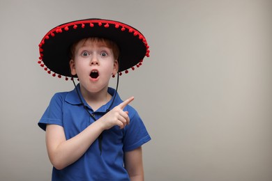 Surprised boy in Mexican sombrero hat pointing at something on grey background, space for text