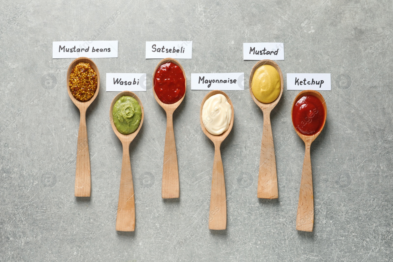 Photo of Different sauces in spoons and name tags on gray background, flat lay