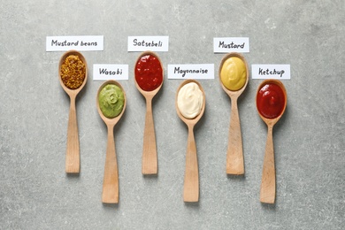 Different sauces in spoons and name tags on gray background, flat lay