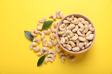 Tasty cashew nuts and green leaves on yellow background, top view