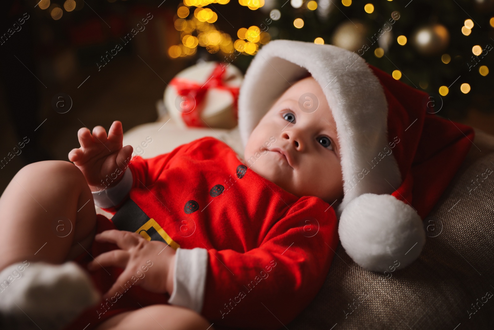 Photo of Cute little baby in Santa Claus suit against blurred festive lights. Christmas celebration