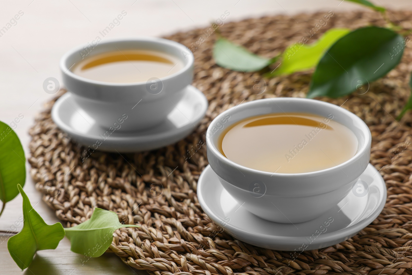 Photo of Green tea in white cups with leaves and wicker mat on table, closeup