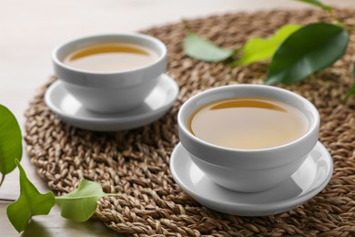 Green tea in white cups with leaves and wicker mat on table, closeup