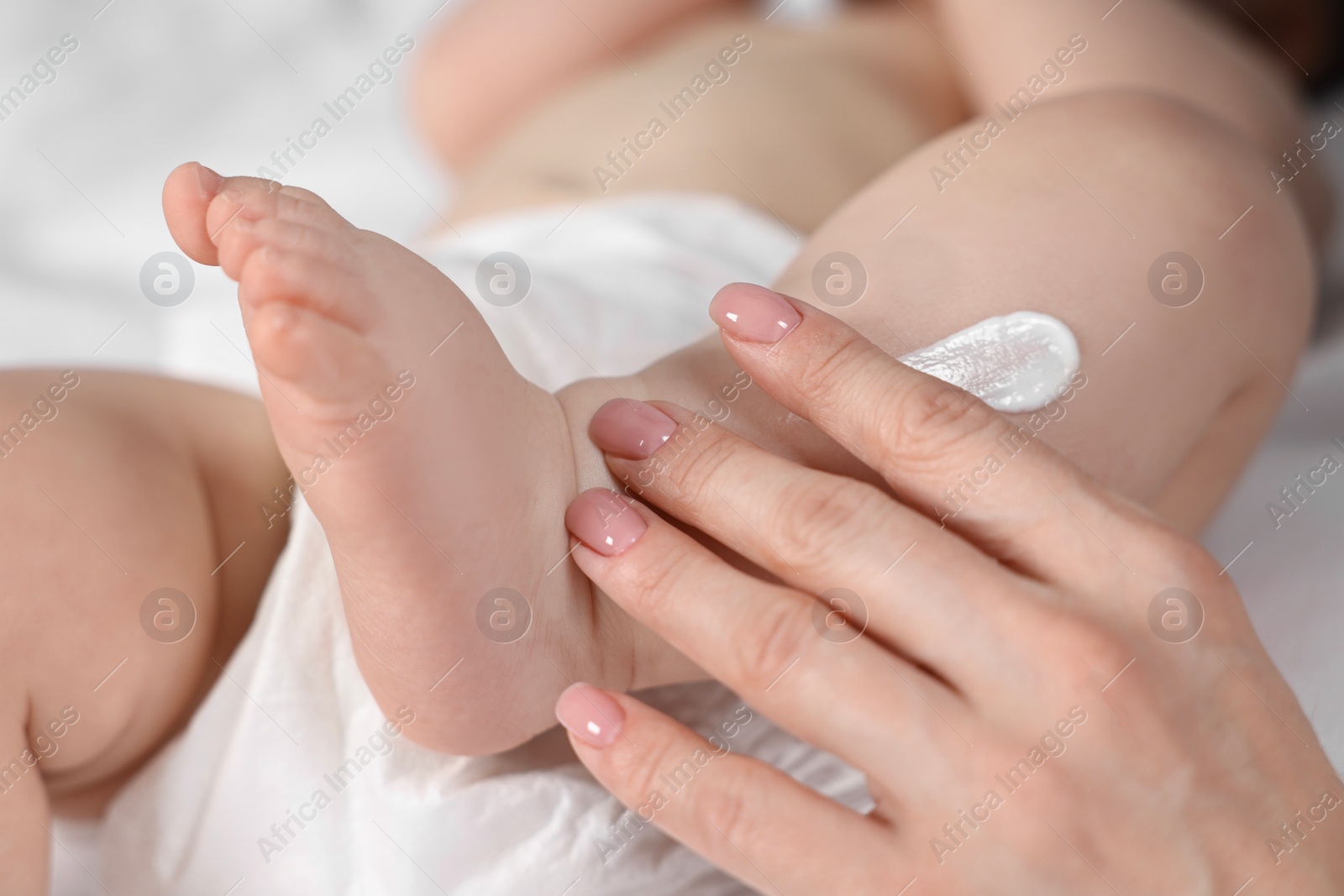 Photo of Woman applying body cream onto baby`s leg on bed, closeup