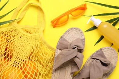 String bag with oranges, sunglasses, cosmetic product, slippers and palm leaves on yellow background, flat lay