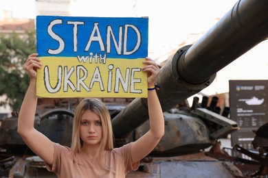 Photo of Sad woman holding poster in colors of national flag with words Stand with Ukraine near broken tank on city street