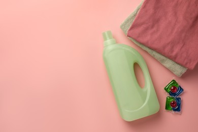 Photo of Bottle of fabric softener, stacked clean clothes and laundry capsules on pink background, flat lay. Space for text