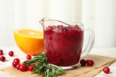 Tasty cranberry sauce in glass pitcher with rosemary and citrus fruit on table