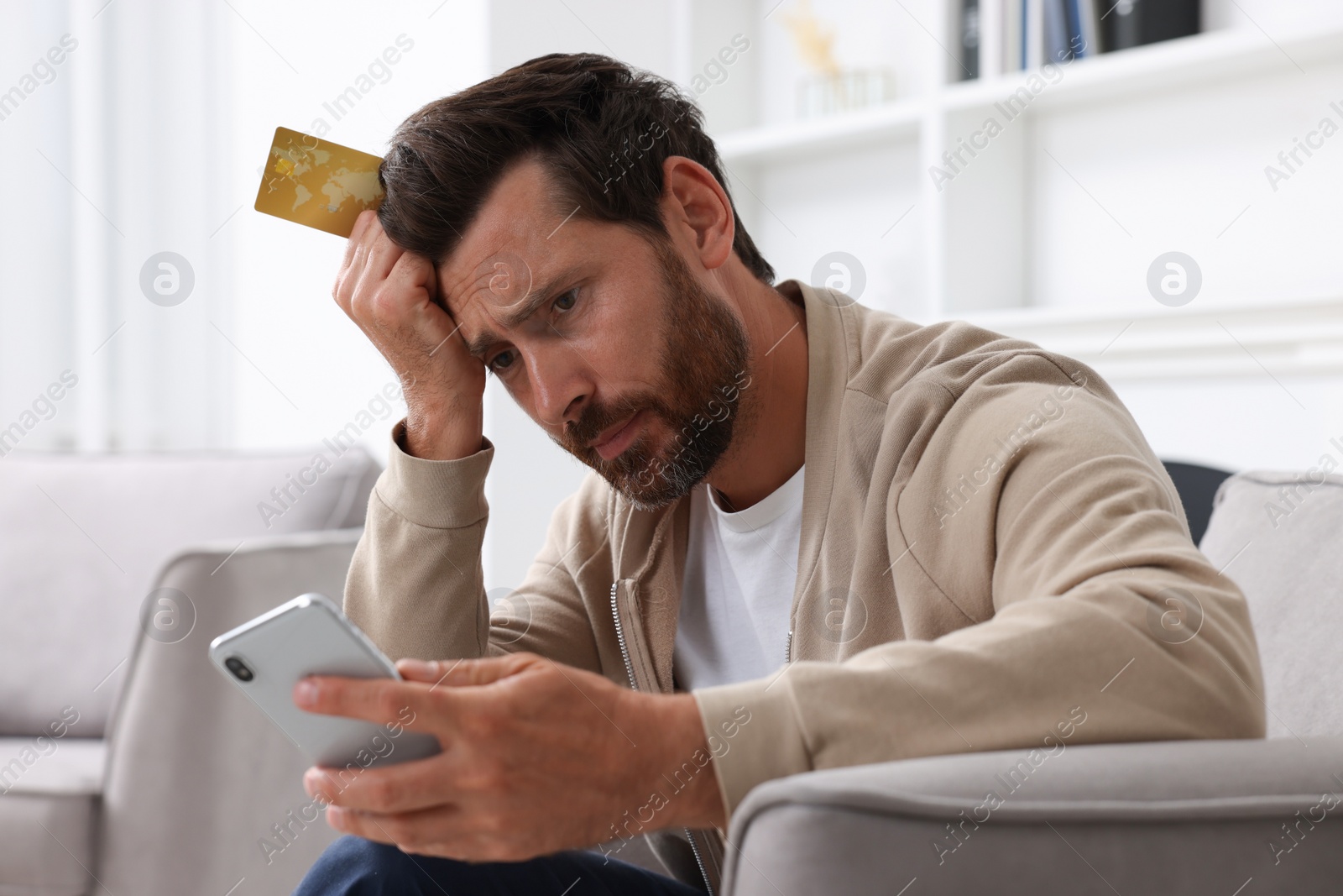 Photo of Upset man with credit card using smartphone in armchair at home. Be careful - fraud