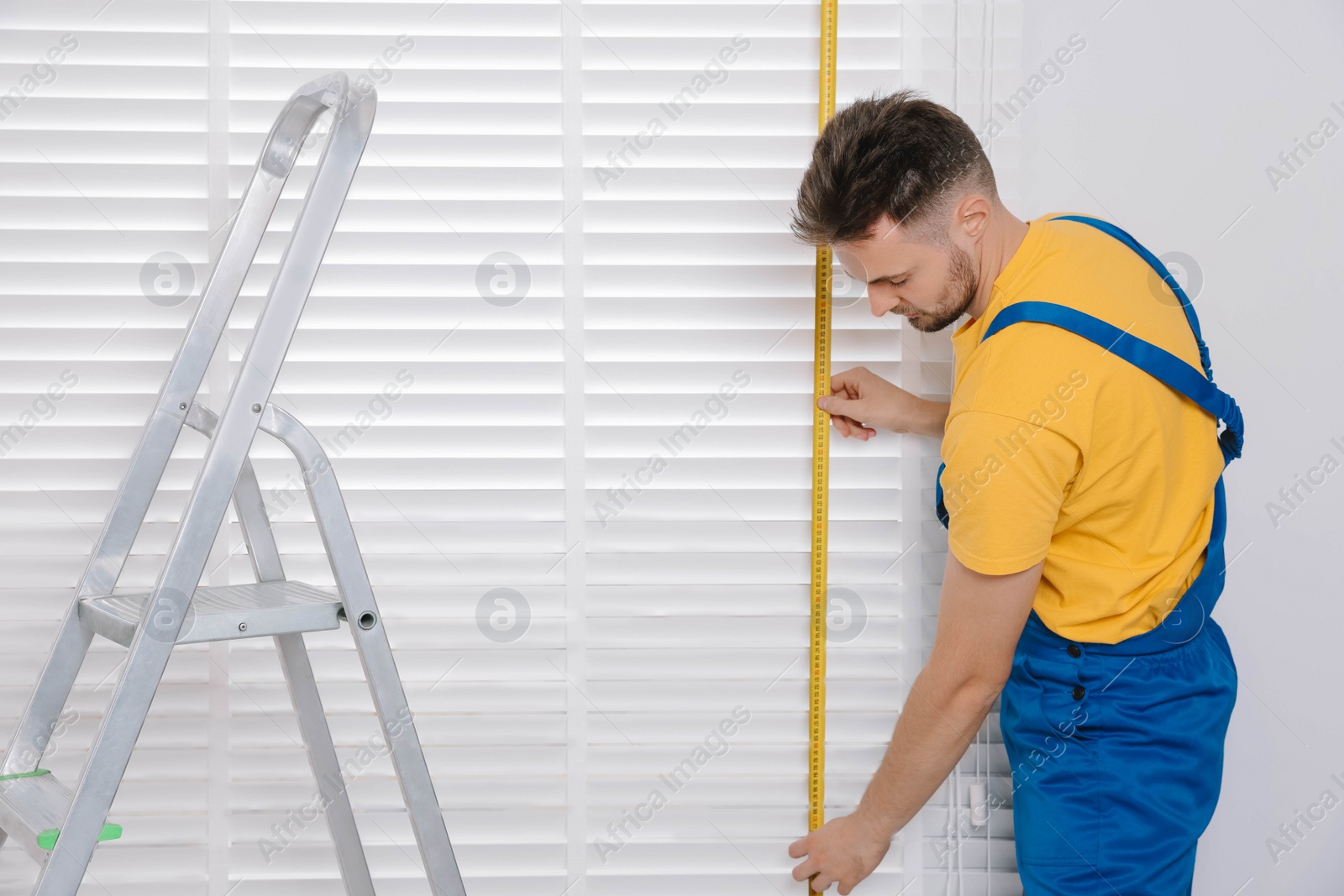 Photo of Worker in uniform using measuring tape while installing horizontal window blinds near stepladder indoors