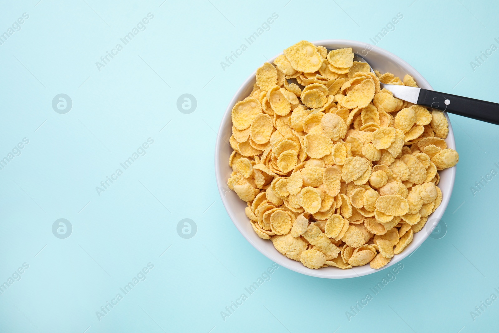 Photo of Breakfast cereal. Tasty corn flakes in bowl and spoon on light blue table, top view. Space for text