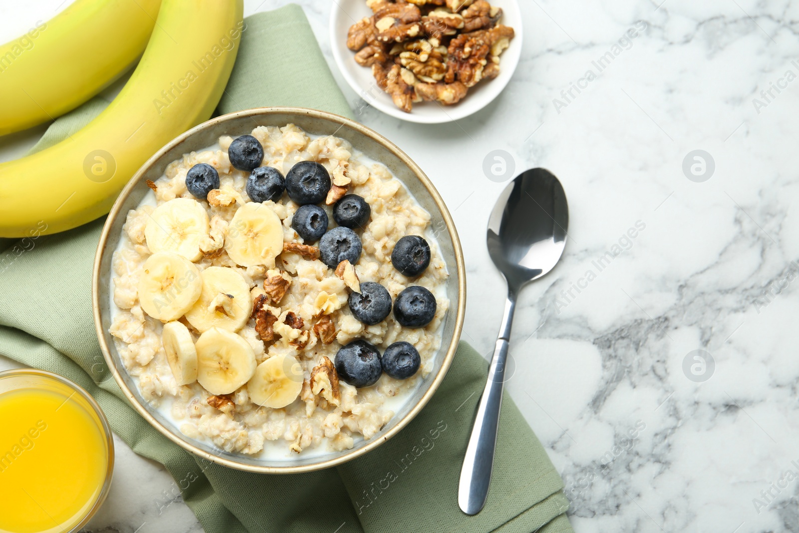 Photo of Tasty oatmeal with banana, blueberries, walnuts and milk served in bowl on white marble table, flat lay. Space for text
