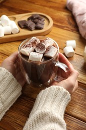 Photo of Woman holding cup of aromatic hot chocolate with marshmallows and cocoa powder at wooden table, closeup