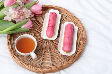 Photo of Tasty breakfast served in bed. Delicious eclairs, tea and flowers on wicker tray, above view