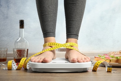 Photo of Woman with measuring tape using scale surrounded by food and alcohol after party on floor. Overweight problem