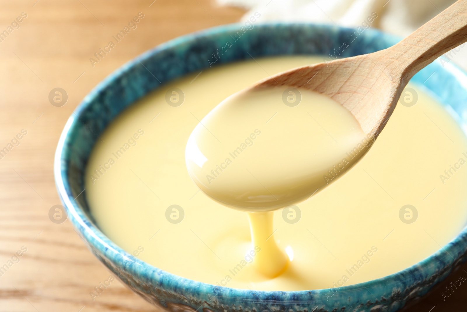 Photo of Spoon of pouring condensed milk over bowl on table, closeup. Dairy products