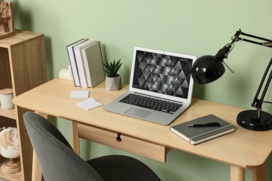 Photo of Modern laptop, books, lamp and stationery on wooden desk near green wall. Home office