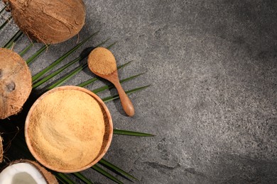 Photo of Coconut sugar in bowl, spoon, palm leaves and fruits on dark textured table, flat lay. Space for text