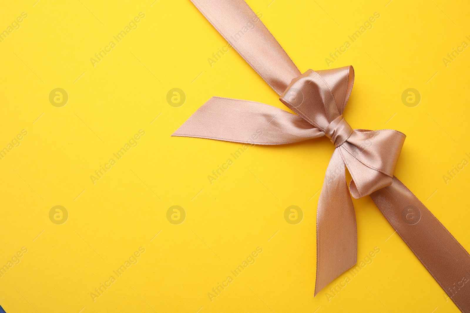 Photo of Beige satin ribbon with bow on yellow background, top view. Space for text