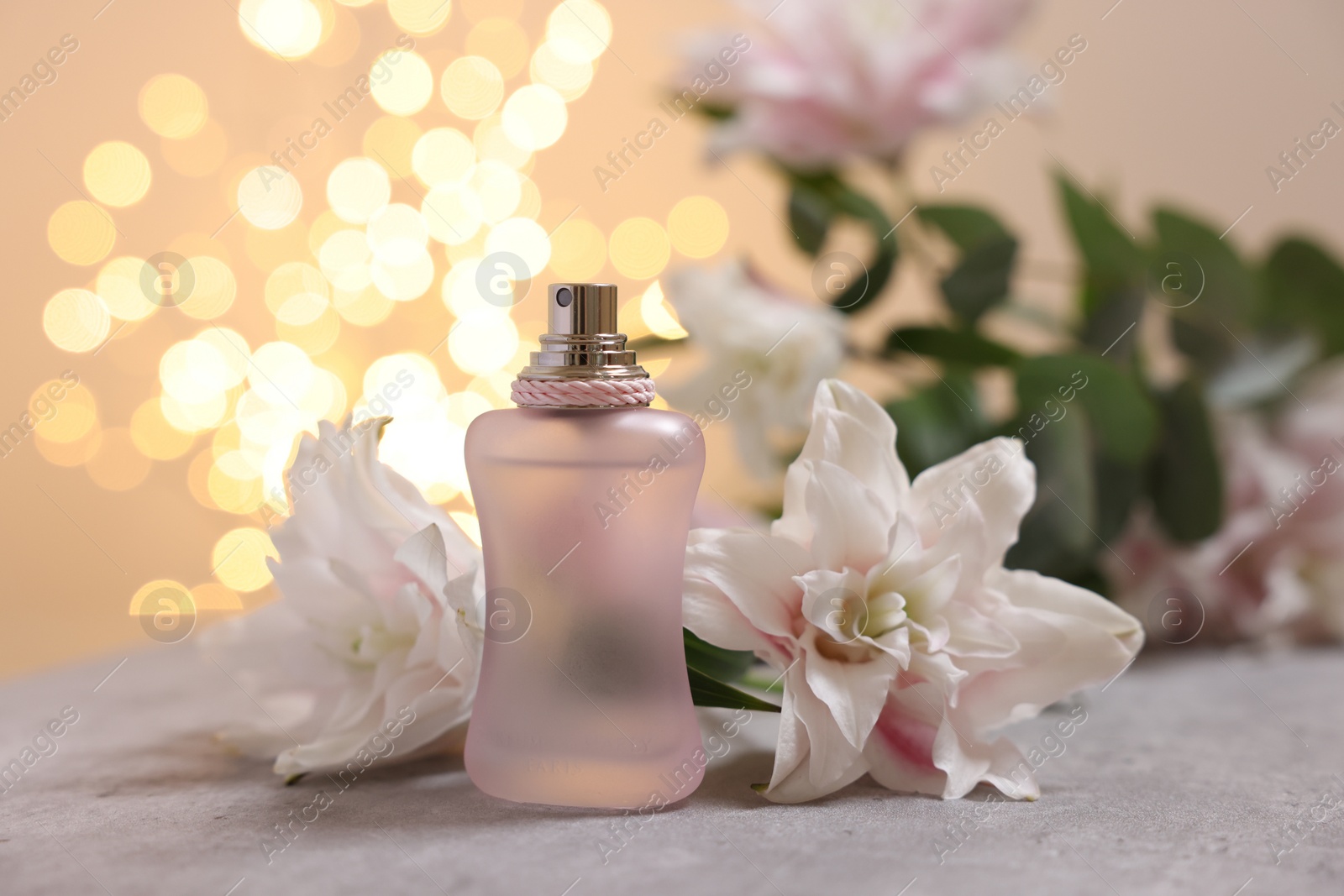 Photo of Bottle of perfume and beautiful lily flowers on table against beige background with blurred lights, closeup