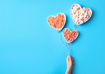 Photo of Woman holding twine near heart shaped bowls full of sweets imitating balloons on light blue background, top view. Space for text