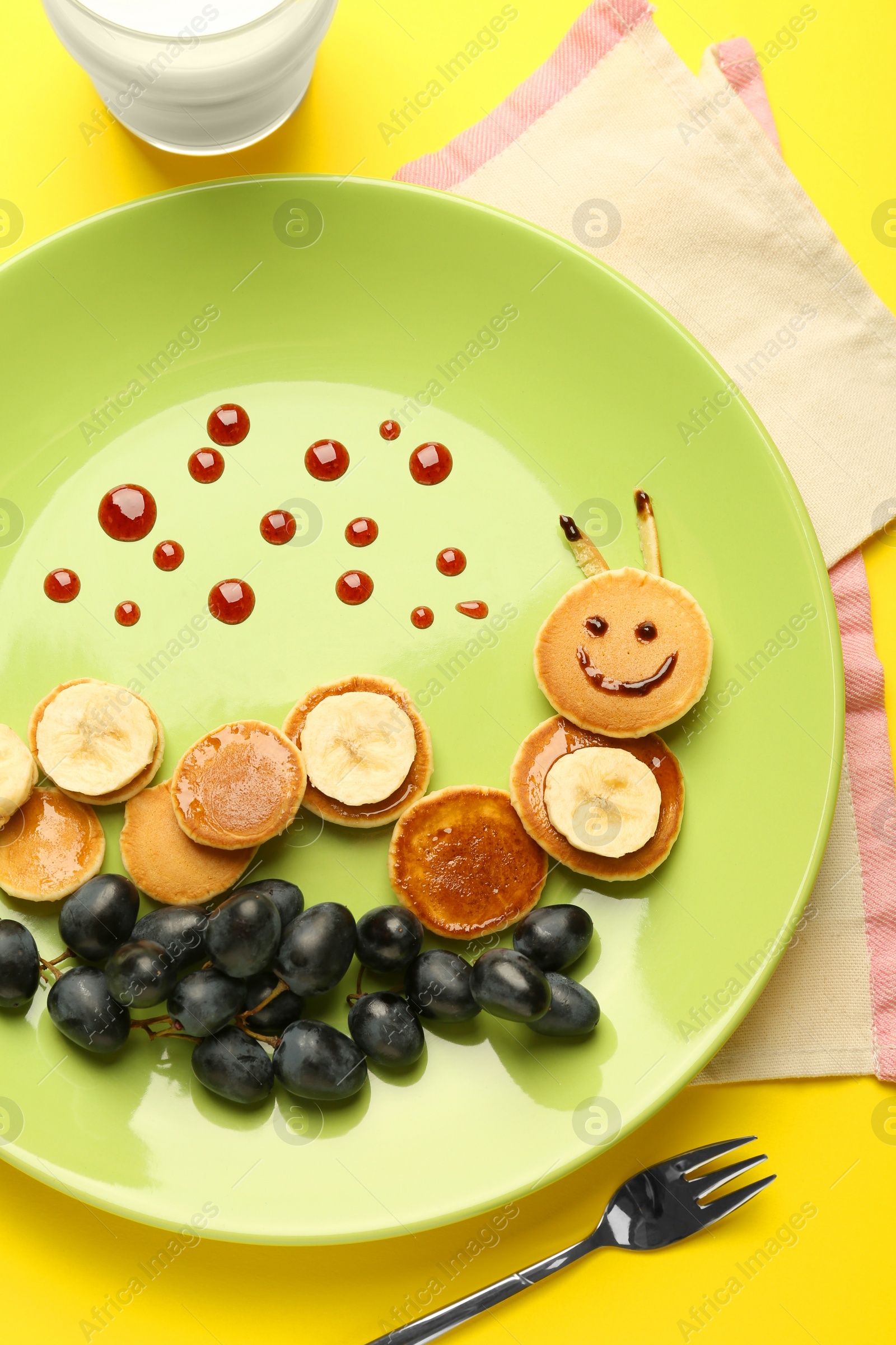 Photo of Creative serving for kids. Plate with cute caterpillar made of pancakes, grapes and banana on yellow background, flat lay