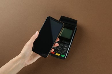 Woman with smartphone using modern payment terminal on brown background, top view