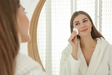 Photo of Young woman with cotton pad cleaning her face near mirror in bathroom