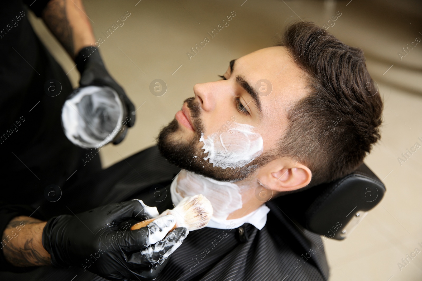 Photo of Professional hairdresser applying shaving foam onto client's skin in barbershop