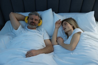 Photo of Smiling man with headphones lying near his snoring wife in bed at home