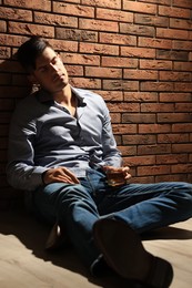 Photo of Addicted man with glass of alcoholic drink sitting on floor near red brick wall