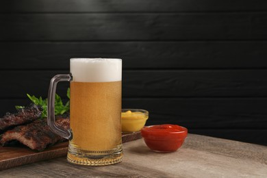 Photo of Mug with beer, delicious grilled ribs and sauces on wooden table. Space for text