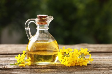 Photo of Rapeseed oil in glass jug and beautiful yellow flowers on wooden table outdoors