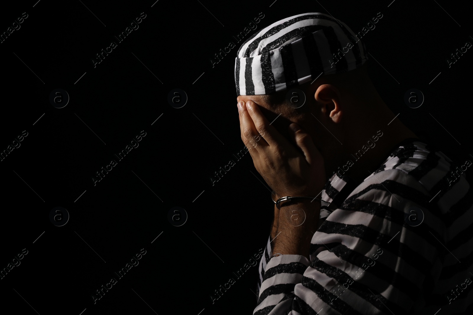 Photo of Remorseful prisoner in striped uniform with handcuffs hiding his face on black background, space for text