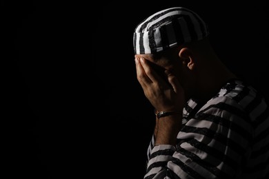 Photo of Remorseful prisoner in striped uniform with handcuffs hiding his face on black background, space for text