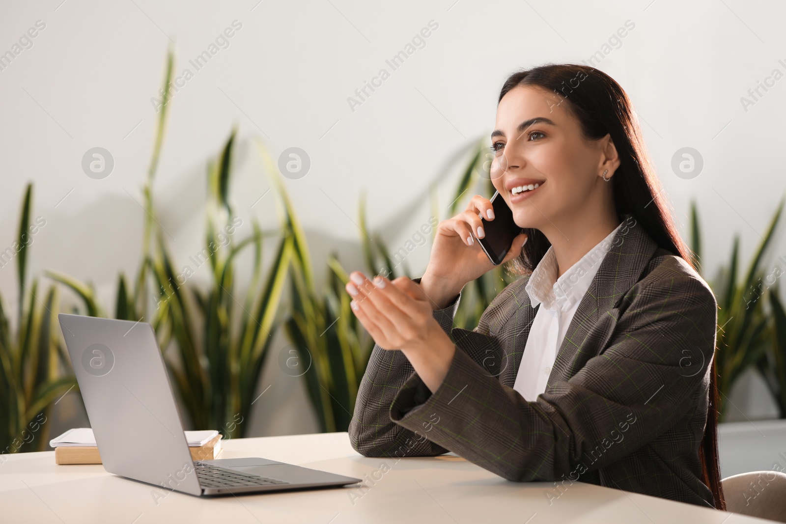 Photo of Happy woman using modern laptop while talking on smartphone at desk in office