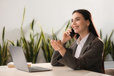 Photo of Happy woman using modern laptop while talking on smartphone at desk in office