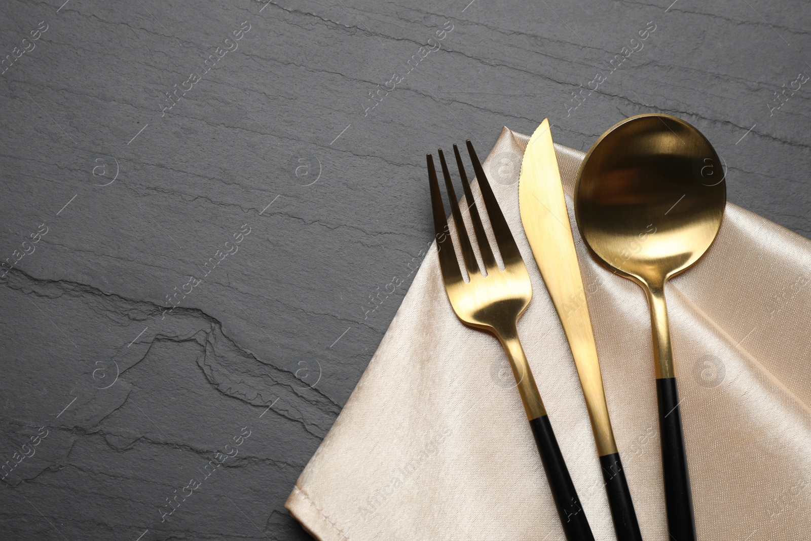 Photo of Set of golden cutlery with napkin on black table, top view. Space for text