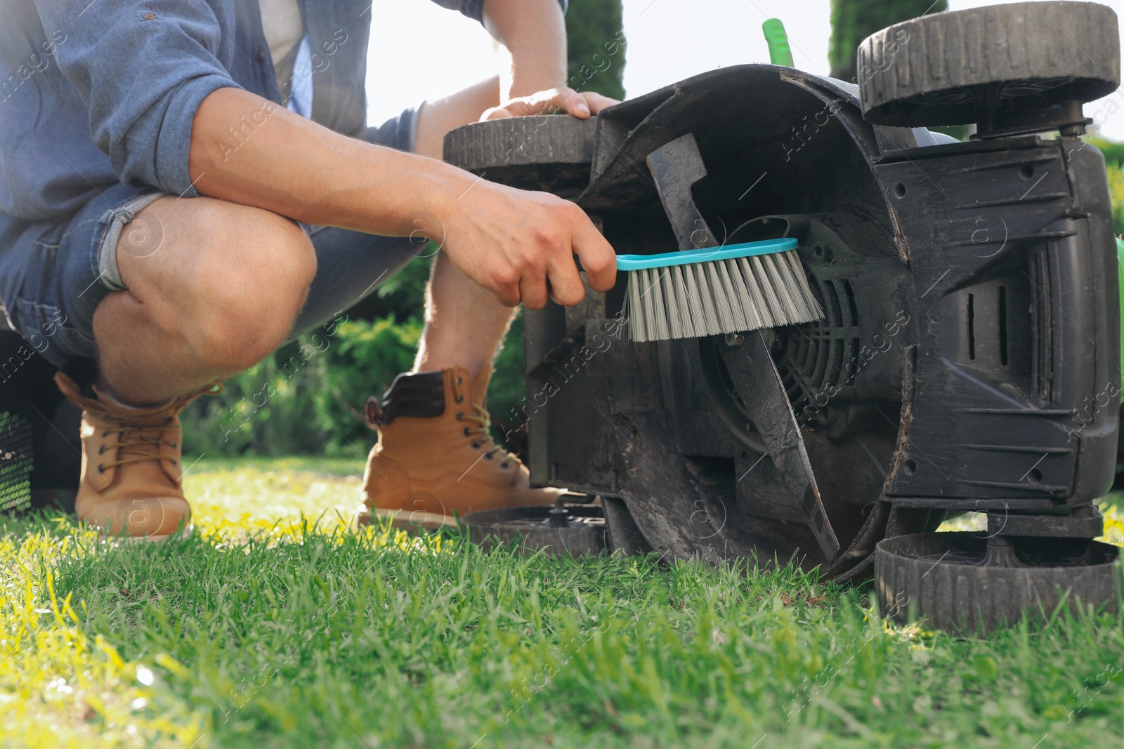 Photo of Young man cleaning lawn mower with brush in garden, closeup