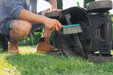 Photo of Young man cleaning lawn mower with brush in garden, closeup