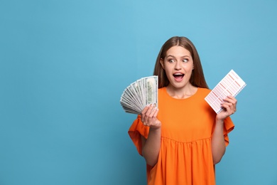 Portrait of emotional young woman with lottery ticket and money fan on light blue background, space for text