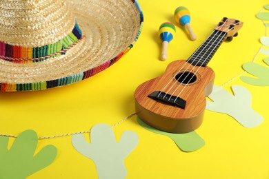 Photo of Mexican sombrero hat, maracas, ukulele and garland on yellow background