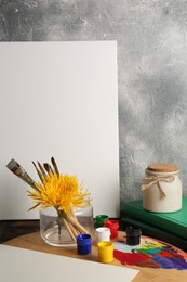 Photo of Blank white canvas, yellow chrysanthemum and different painting tools on wooden table