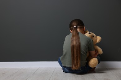 Photo of Child abuse. Upset girl with toy sitting on floor near grey wall, back view. Space for text
