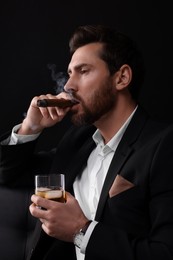 Photo of Handsome man in elegant suit with glasswhiskey smoking cigar on black background