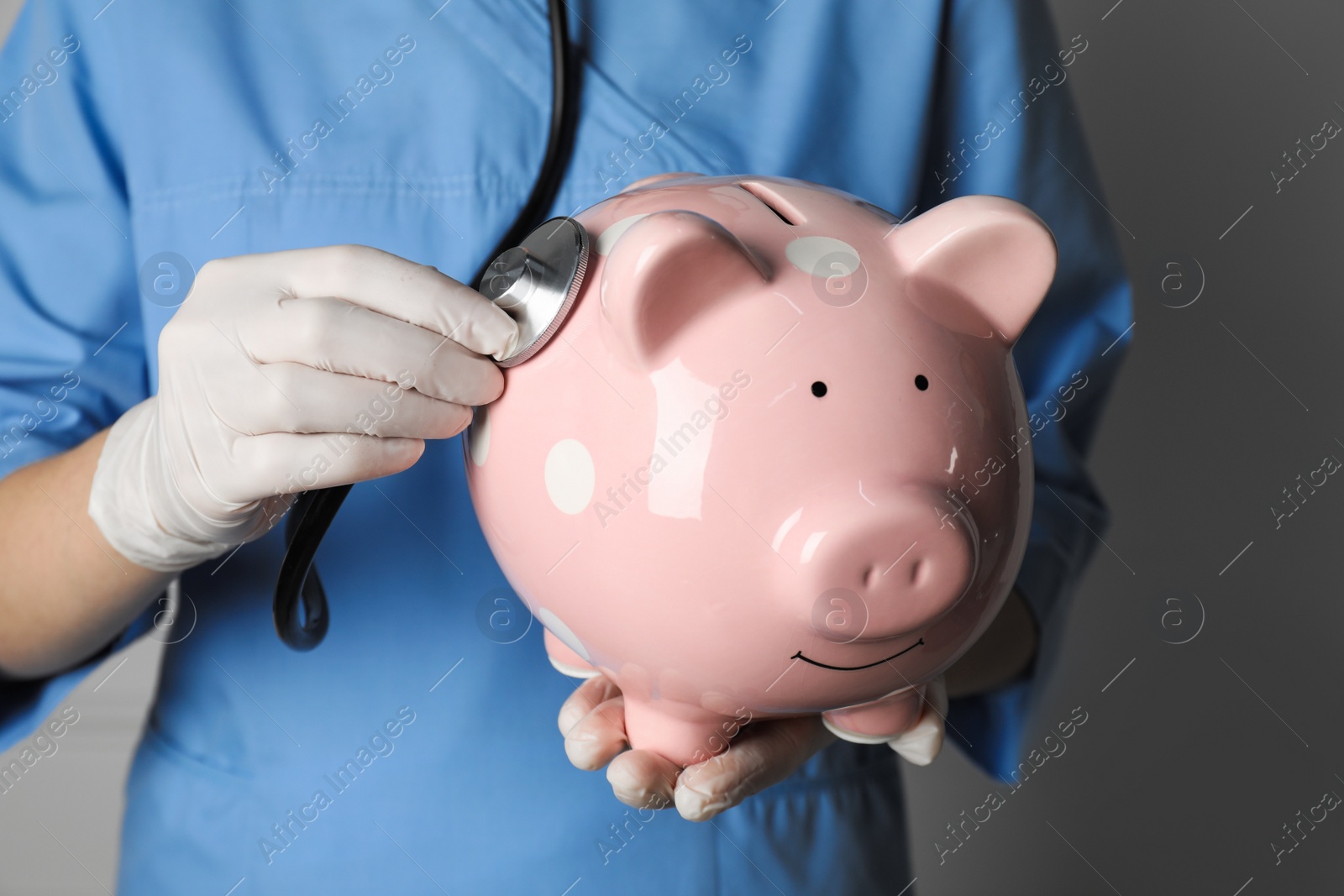 Photo of Doctor holding pale pink ceramic piggy bank with stethoscope against white wall, closeup. Medical insurance