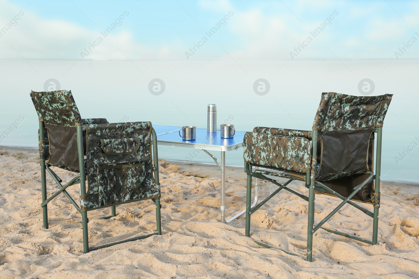 Photo of Camouflage fishing chairs and table with metal cups on sandy beach near river